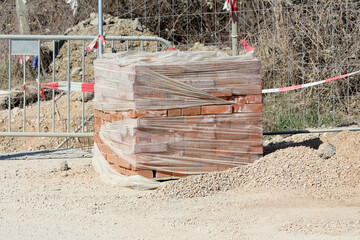 A large stack of red clay bricks is tightly wrapped in clear plastic film, resting on a...