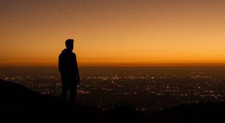 Fototapeta premium Silhouette of a man standing on a hill overlooking a city at sunset, with a warm orange sky.