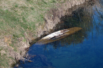 An old wooden boat lies overturned and partially submerged in transparent, shallow water near a grassy bank, its hull visibly weathered and marked by years of exposure