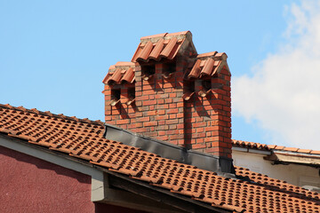 A sturdy brick chimney with three terracotta tile caps rises above a red clay tiled roof,...