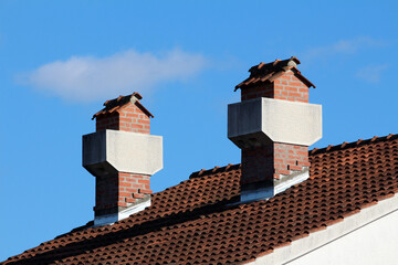 Two brick chimneys with distinctive concrete hoods rise from a steep terracotta-tiled roof, their angular tops and red brickwork contrasting against a vivid blue sky