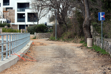 A rough gravel road under construction ends at a blue dead end sign, bordered by bare trees and modern white apartments, with scattered construction debris along the seaside path