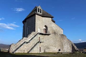 Fototapeta premium A small medieval church with a bell gable and steep roof sits atop a grassy hill, surrounded by a partially ruined stone wall and metal stairs, overlooking a rural landscape under blue sky