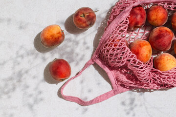 Fresh peaches in a pink string bag on white sunlit background with shadow. Healthy food concept. Eco friendly theme. Summer backdrop