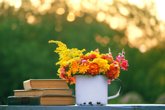 white enamel mug with colorful flowers and books on table in garden close up. summer nature background. romantic rustic floral composition with seasonal flowers
