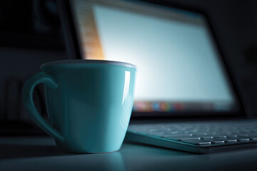 Blue ceramic cup, keyboard, and blurred laptop screen create cozy night workspace atmosphere, illuminated by soft blue light, evoking calm and focus for late work sessions