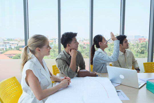 Diverse business team discussing project in bright meeting room with blueprints and laptop, symbolizing teamwork, innovation, and modern corporate collaboration.