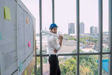 Caucasian architect wearing a safety helmet reviewing large blueprints in a modern office with panoramic city view, lit by bright natural sunlight.