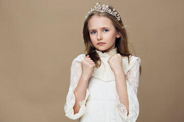 Young girl in a white dress with a crown, looking serious and elegant against a soft beige...