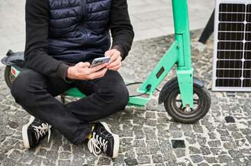 Cropped view of man sits on electric scooter with solar panel and smartphone while charging....