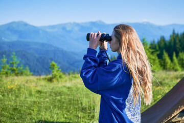Woman stands in grassy meadow, using binoculars to observe distant mountains, wearing in blue...