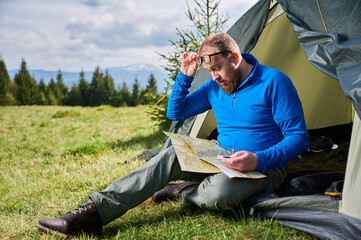 Man sits in tourist tent's entrance, deeply focused on map. Tourist holds compass and contemplates...