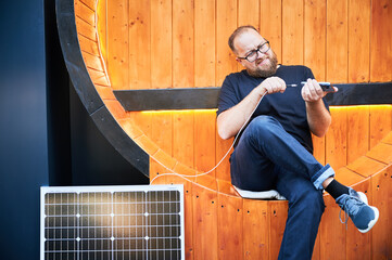 Bearded man connecting smartphone to photovoltaic solar panel. Integration of sustainable renewable...