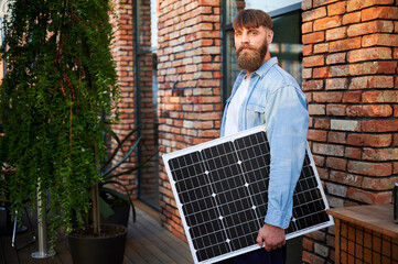 Portrait of bearded man holds solar panel, standing in front of brick building and lush greenery....
