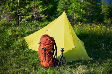 Yellow tourist tent on grassy hillside, accompanied by orange backpack and camera tripod.
