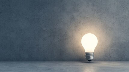 A glowing light bulb stands on a concrete surface against a textured gray wall, symbolizing ideas, innovation, or creativity.