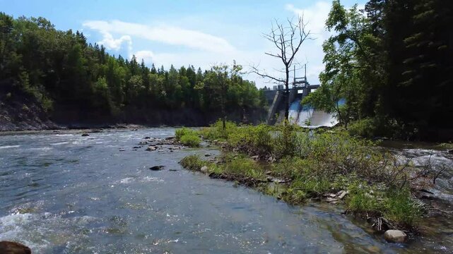 Aerial drone footage moving above the Mitis River through riverside greenery toward the fortified appearance of Mitis-2 Dam, a concrete hydroelectric structure in Price, Quebec, Canada.