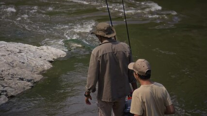 Rear follow shot of African American grandfather and biracial grandson hiking along stony river bank, carrying tackle, rods, gear box, searching for fishing spot and talking, on sunny summer day - Powered by Adobe