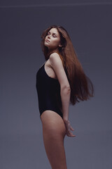 young woman wearing black bodysuit stands sideways with eyes closed and long hair flowing against...