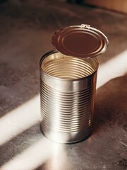 Opened silver metal can with lid still attached, placed on cracked light-gray surface with sunlight and shadows, evoking raw domestic contrast