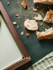 Wooden tray with scattered breadcrumbs and bread bits beside white plate and green checkered cloth — evoking rustic everyday aftermath
