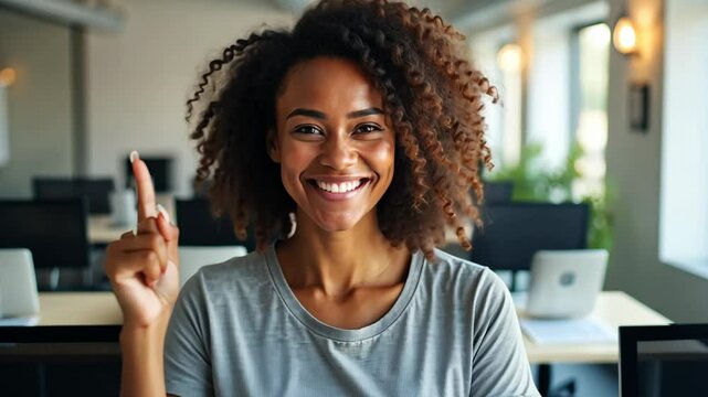Happy optimistic woman in casual t-shirt standing in modern office environment, her arm raised in fist, successful finish of difficult work. She surrounded by computers and open space elements.