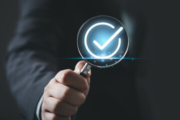 A hand holding a magnifying glass with a checkmark symbol, representing verification, approval, or...