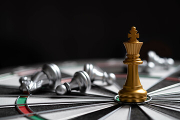 A golden chess king stands prominently on a dartboard, surrounded by silver chess pieces,...