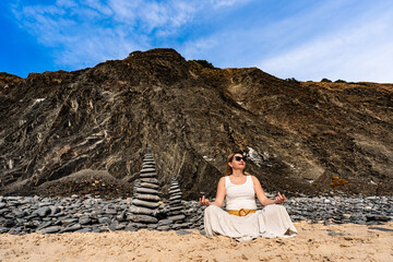Beautiful mid adult woman meditating on sandy beach against dark cliff background on summer day