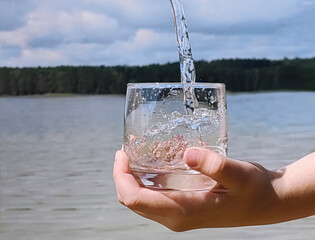 a man holds a glass of clear water in his hand against the backdrop of a forest lake