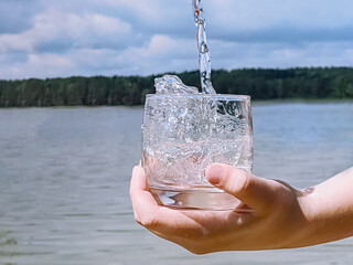 a man holds a glass of clear water in his hand against the backdrop of a forest lake