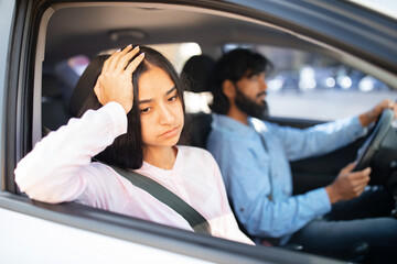 Sad Indian woman looking out car window with hand on head, while man driving silently. Displays...