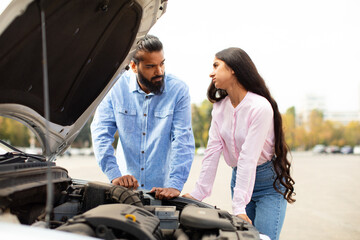 Indian couple leaning on broken cars hood, having serious conversation. They both look concerned,...