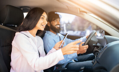 Cheerful Indian woman pointing out window with phone while man driving car. Represents navigation, excitement, and teamwork during fun road trip moment, closeup