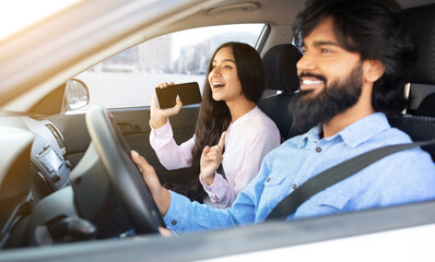 Indian woman singing with smartphone while her boyfriend driving car. Cheerful spouses expressing fun, discovery, and tech savvy moment during joyful car ride, closeup