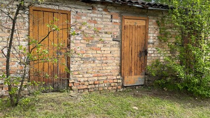 A weathered brick building with wooden doors is situated amidst greenery.