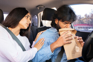 Worried Indian woman supports anxious man breathing into paper bag while driving. Scene of...