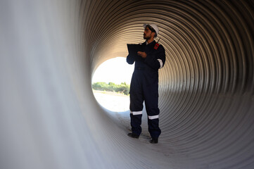 A male mechanical engineer in safety uniform is inspecting and examining a piping system for a new construction project of a large industrial building.