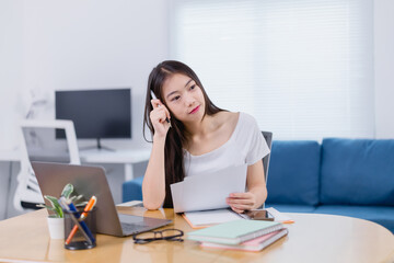 Young Asian businesswoman working from home, sitting at a desk with a laptop and documents, holding...
