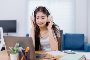 Young Asian student wearing headphones, listening to music while studying online with a laptop at...