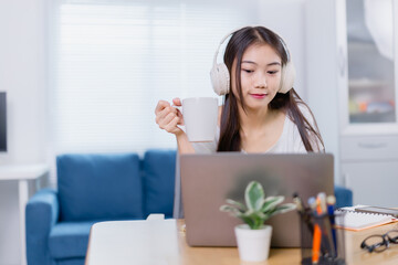 Asian freelancer working from home drinking coffee and wearing headphones while using her laptop in her living room, in a cozy work from home setup