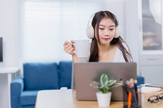 Asian freelancer working from home drinking coffee and wearing headphones while using her laptop in her living room, in a cozy work from home setup