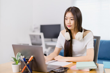 Concentrated young Asian woman wearing headphones around her neck and taking notes in notebook...