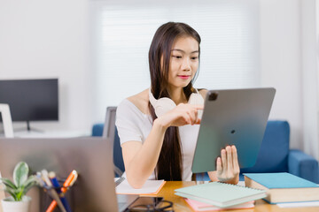 Young asian woman wearing headphones using tablet and studying online at home office with books and...