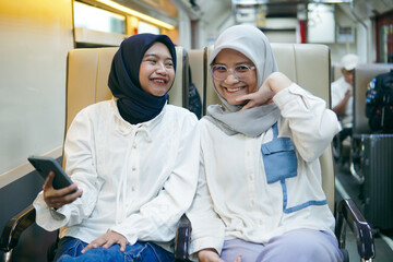 Two Asian hijab woman chatting and smiling while using smartphones on train seats, enjoying their journey together