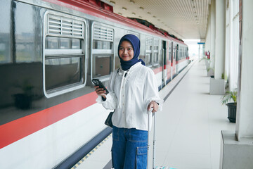 Young Muslim woman in hijab standing at train station platform with suitcase and phone, smiling confidently while waiting for departure