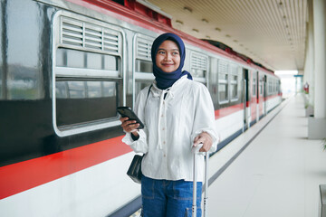 Young Muslim woman in hijab standing at train station platform with suitcase and phone, smiling...
