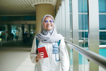 Asian woman in hijab walking through airport corridor, holding her passport and heading confidently toward departure gate
