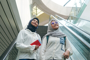 Two Asian women in hijab riding an escalator at the airport, carrying bags and heading toward their departure gate