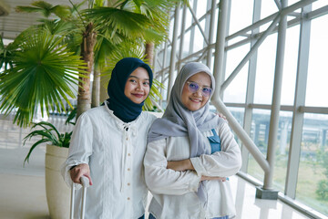 Two Asian women in hijab chatting cheerfully at the airport, enjoying their time together before...